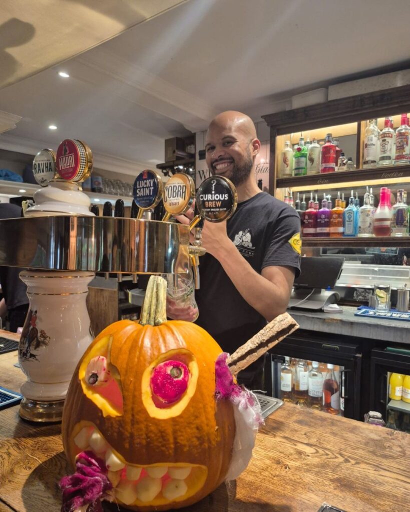 xarved pumpkin sat on a bar with bartender pouring a pint of larger in a country pub